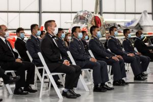 Photo of graduates in military dress sitting on white chairs in an aircraft hangar.