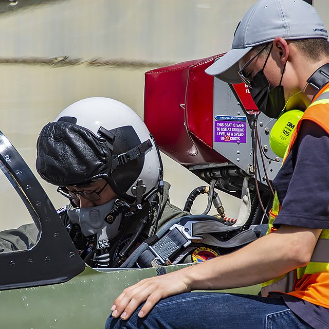 itps20216290069 Fighter pilot in green flight suit sitting in an Aero Vodochody aircraft. ITTC flightline crew member is shown in safety vest.